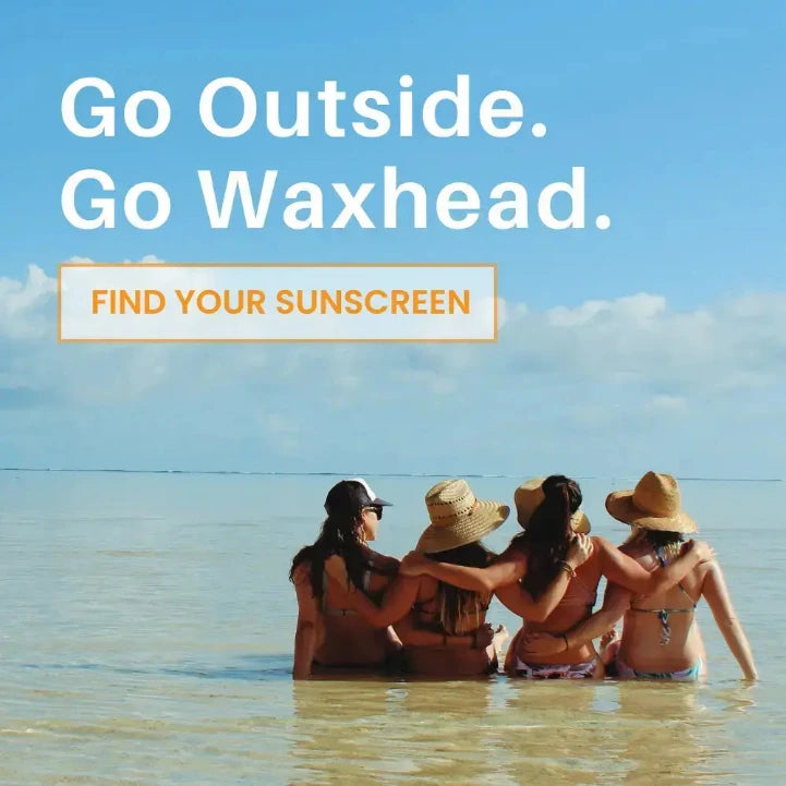 Four women sitting in shallow ocean water facing the sea under a blue sky, with large text reading 'Go Outside. Go Waxhead.